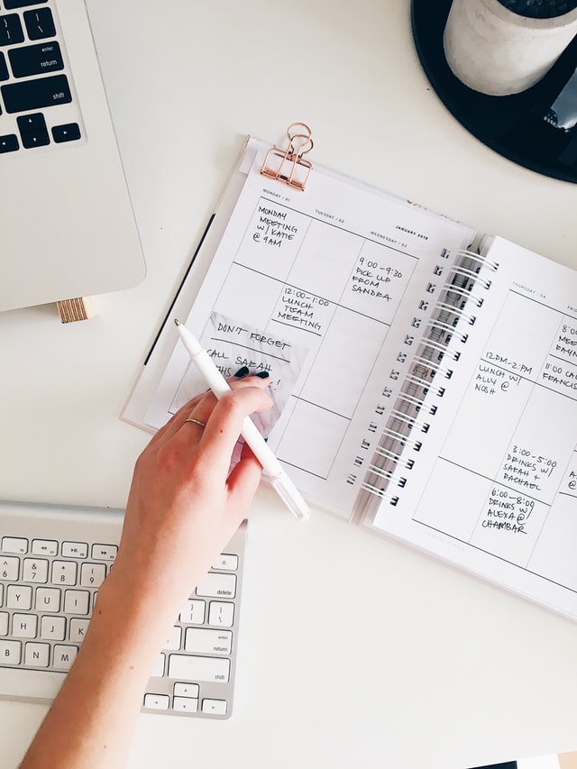 overlay shot of a planner filled out next to a laptop with a hand reached out touching it with a pen in hand