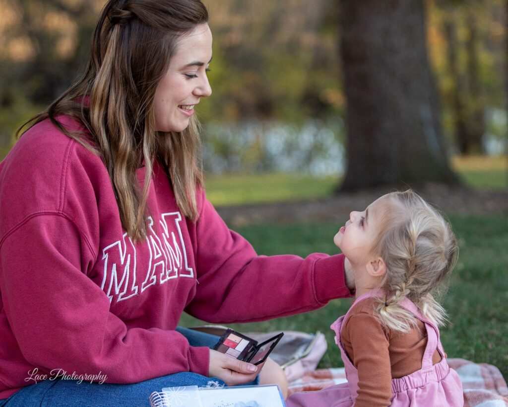 Hannah Christine sitting on a picnic blanket in the grass facing her daughter, smiling, putting makeup on her. Her daughter is smiling up at her. Hannah does color analysis, branding and life coaching in Lake Country, WI