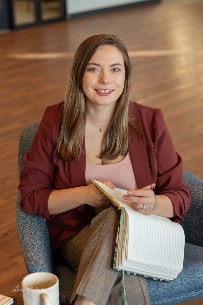 Headshot of Hannah wearing a merlot colored blazer and plaid pants, sitting in a chair with a notebook out, Hannah is a life and wellness coach and branding specialist for Lake Country, Oconomowoc, WI