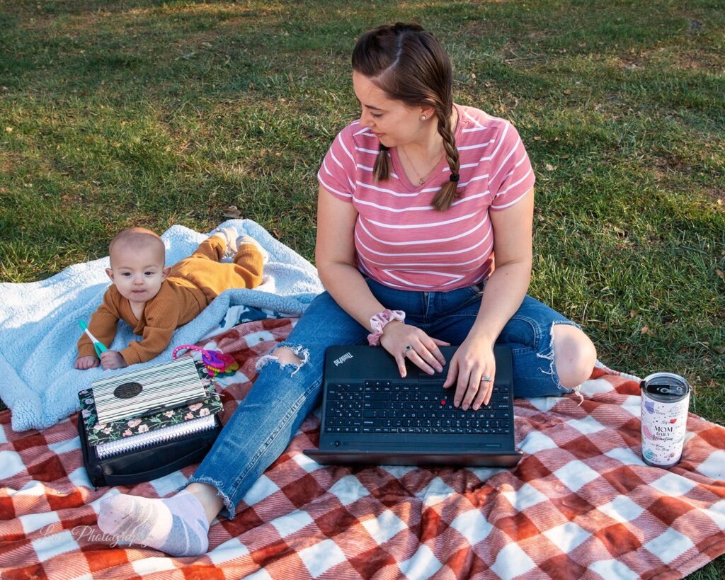 Hannah sitting on a checkered picnic blanket in the grass with her laptop out, looking to her right at her baby son laying on the blanket next to her. Hannah is a life and wellness coach in Oconomowoc, WI