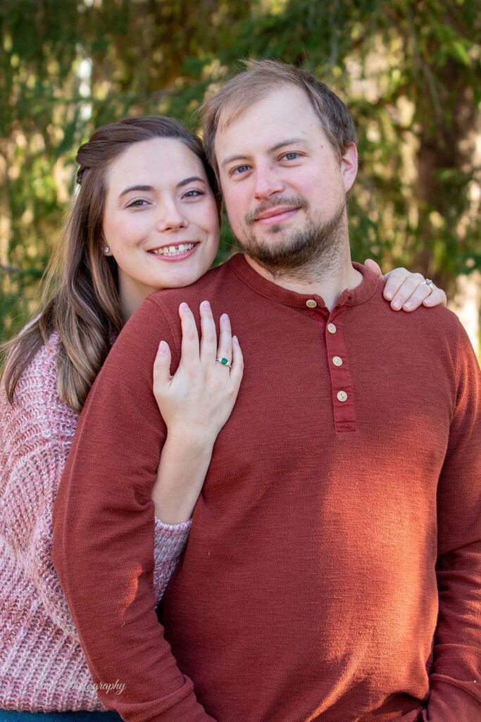 Hannah posing with her husband outdoors in front of a tree, smiling with her arm linked under his arm and hand on his shoulder