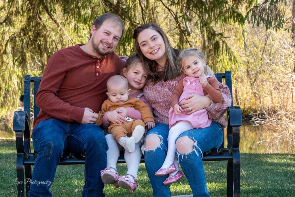 Hannah and her beautiful family: husband, two daughters and baby son sitting on a bench hugging smiling at the camera outdoors with a tree behind them. Hannah owns a family-friendly branding and color analysis business helping moms and professionals in Waukesha, WI