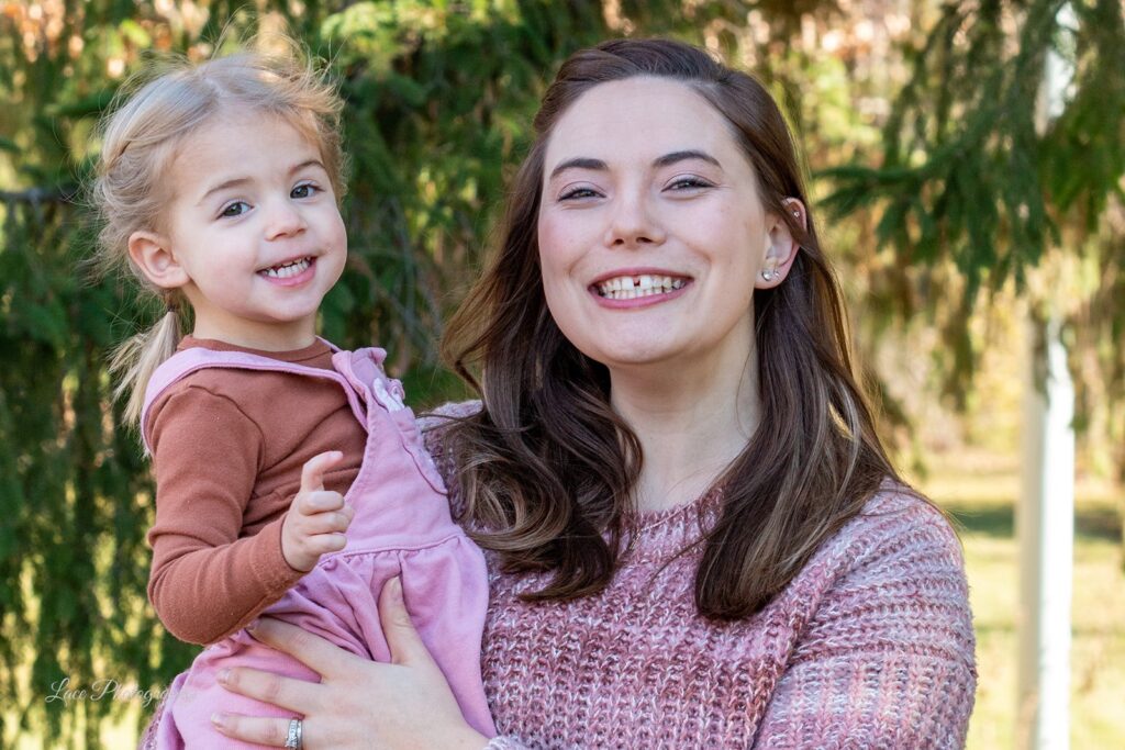 Hannah holding her daughter on her hip, both are smiling wide, looking at the camera, posing outdoors in front of a tree in the sunshine. Hannah is a wellness and life coach in Lake Country, WI