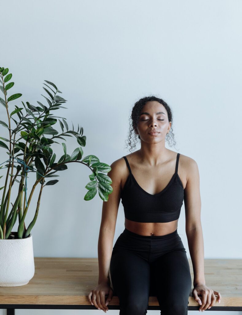 Girl in a black workout set closing her eyes, meditating next to a green plant