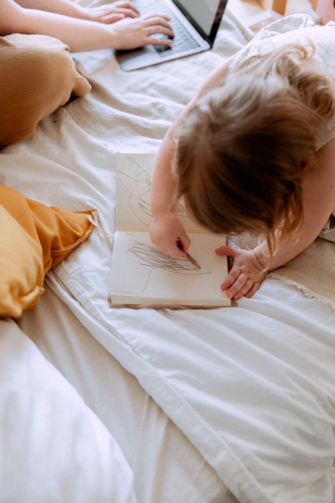 From above of small girl in dress drawing with pencil in notebook while mother using laptop on bed near